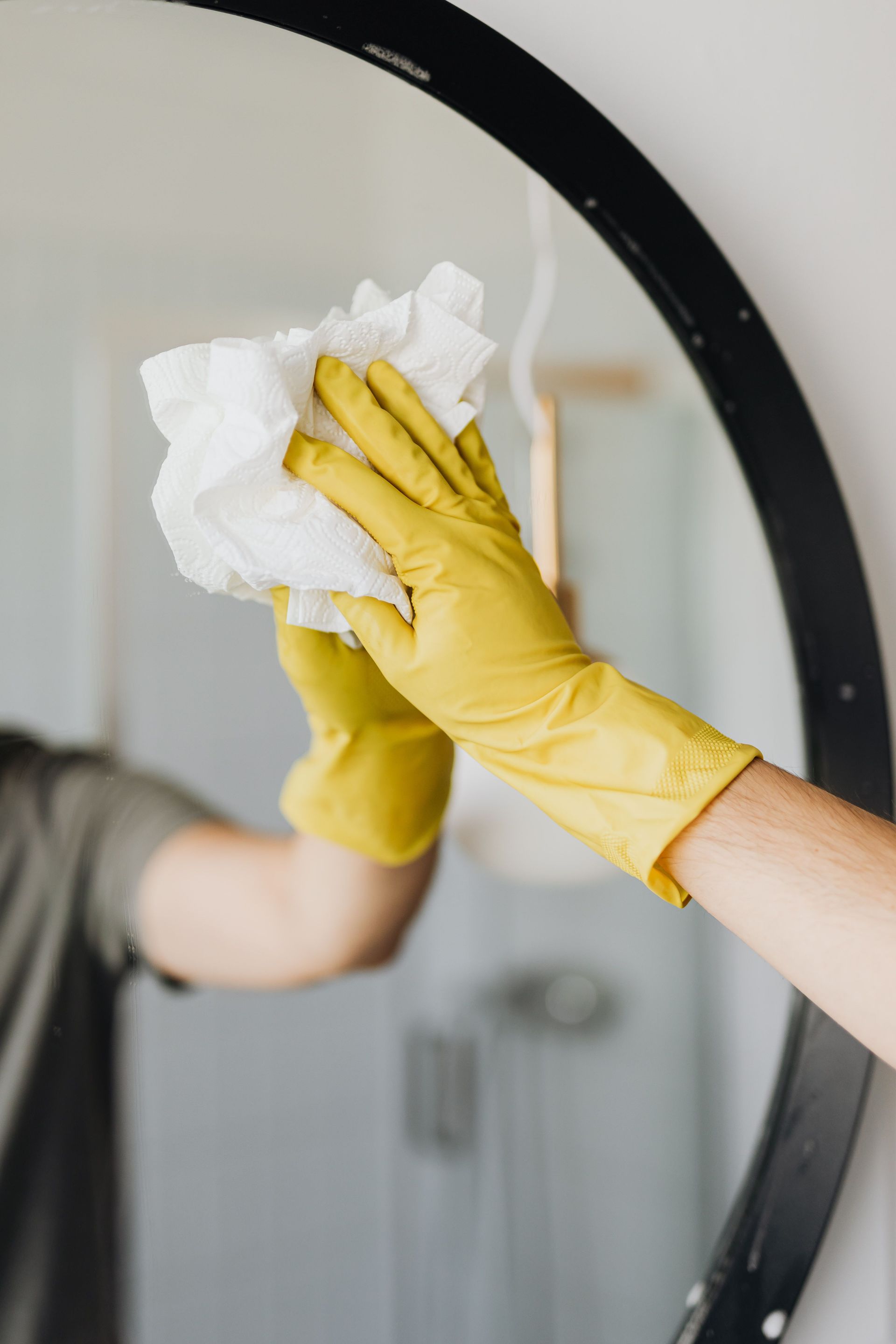 Crop male in yellow latex glove wiping mirror with paper napkin and detergent in bathroom while standing and working at home